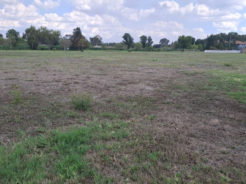 A wide, level vacant residential plot featuring open grassland with surrounding mature trees and nearby residential development.