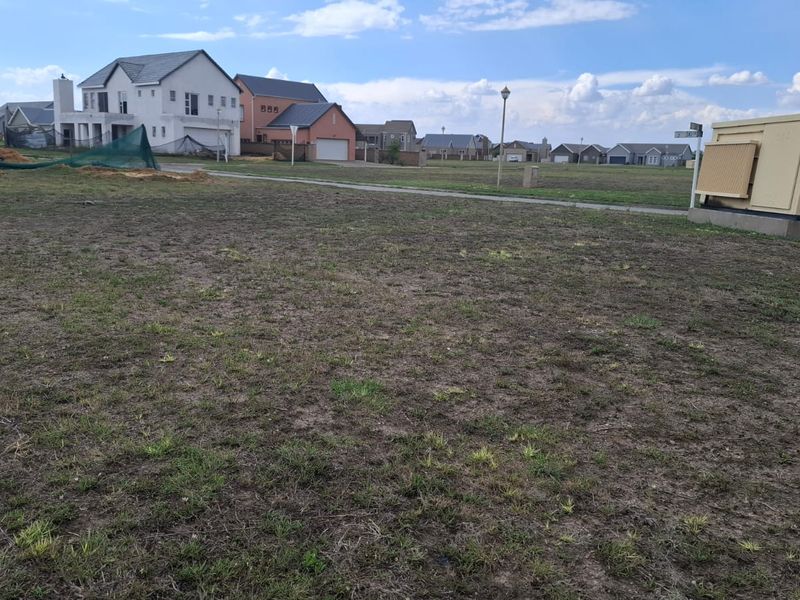 A vacant residential plot featuring sparse grass cover, an electrical transformer box, and surrounding suburban housing in a developed estate.
