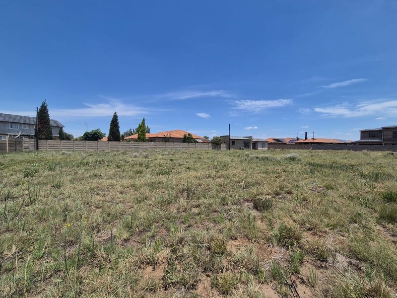 A vacant residential plot featuring natural grass cover and enclosed by a precast concrete boundary wall in a suburban neighborhood.