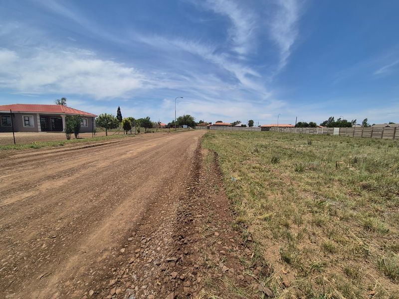 A view of an unpaved residential street bordering a vacant plot of land with a precast concrete boundary wall in the background.