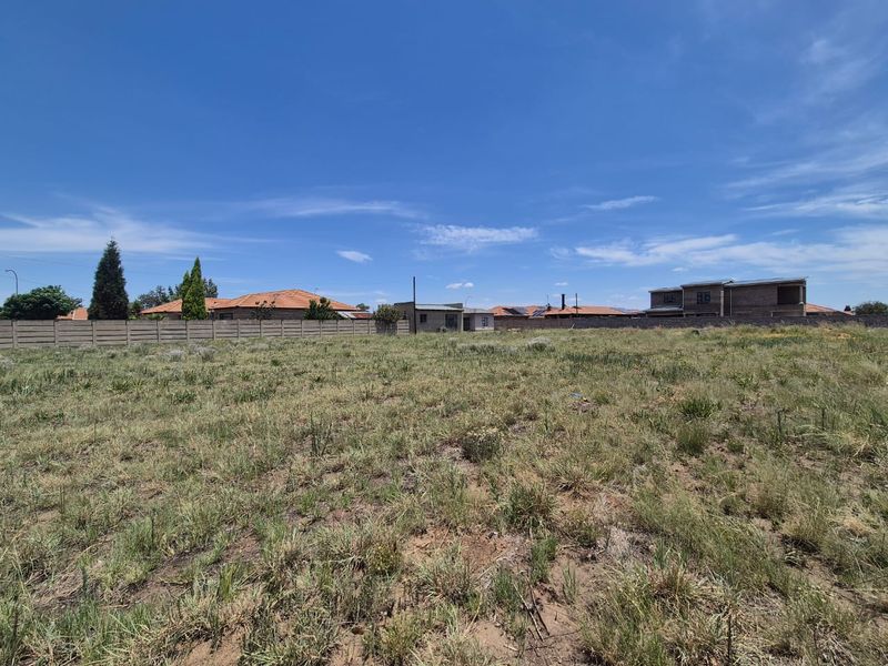 A view of a vacant residential plot featuring natural grass cover and a perimeter wall, situated in a developed neighborhood.