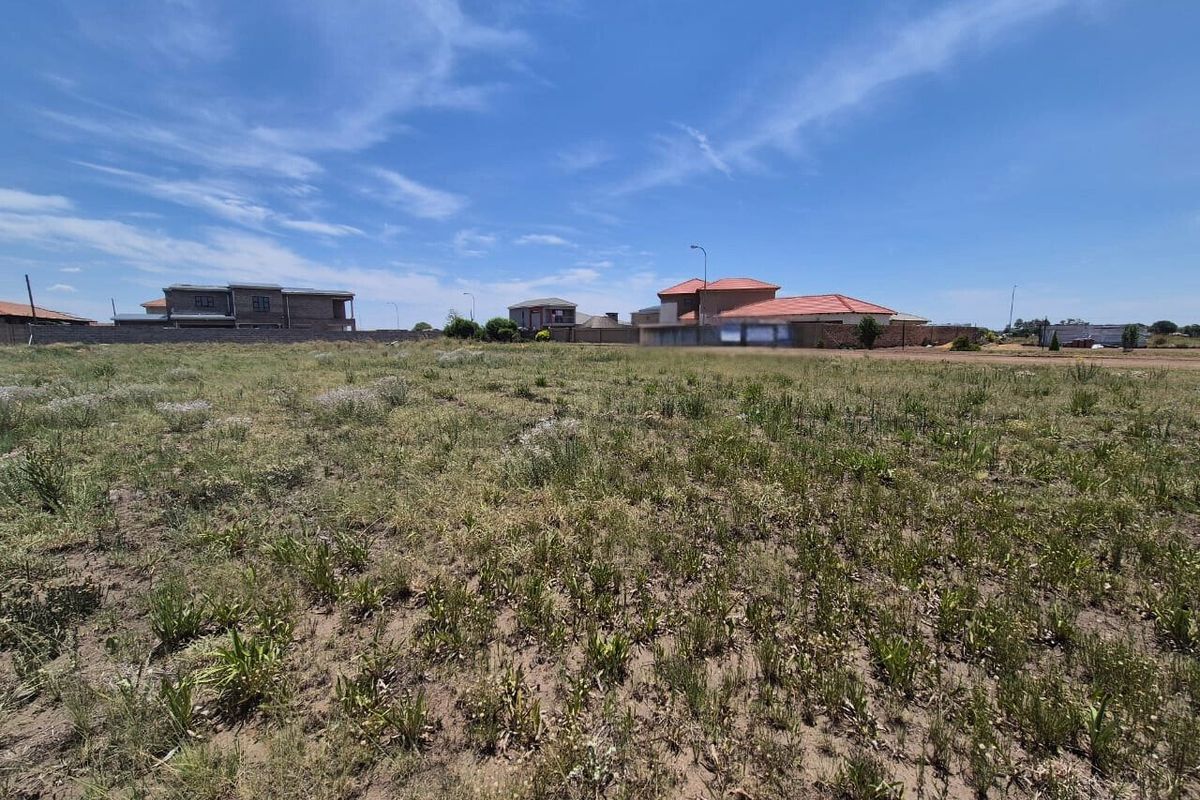 A view of a vacant residential plot of land with surrounding boundary walls and neighboring houses in the distance.