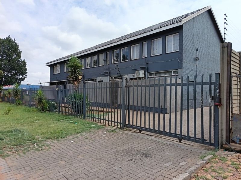 Exterior view of a multi-story flat complex featuring a dark painted brick facade, tiled roof, and secure metal perimeter fencing with a paved driveway.