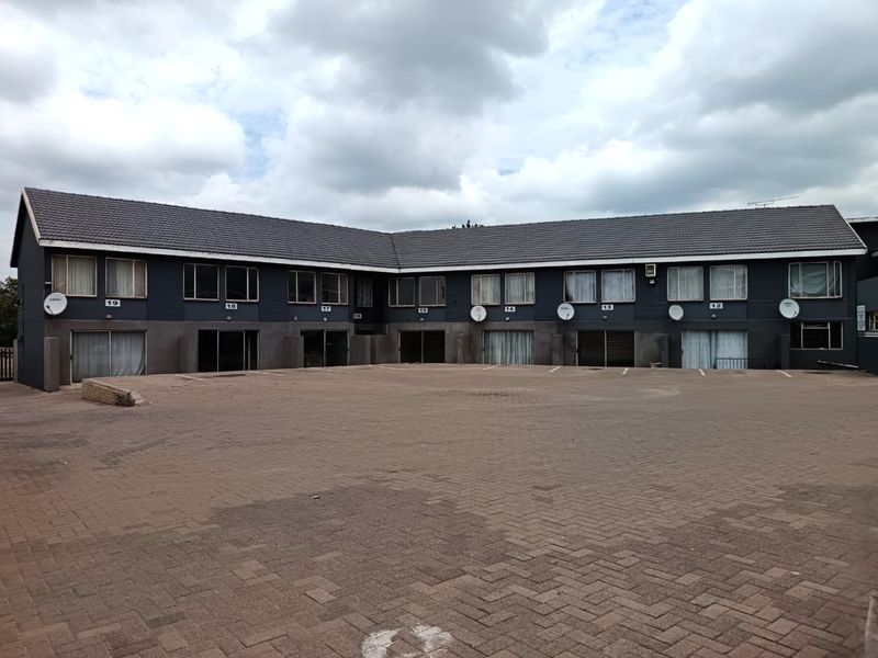 Exterior view of a multi-unit residential flat complex featuring a large paved parking area and dark-toned facade with multiple ground-floor sliding door entrances.