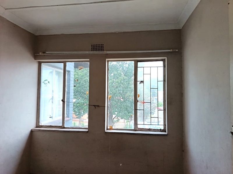 An empty bedroom featuring dual windows with security bars, ventilation grille, and curtain rail, requiring cosmetic updates to the walls and window frames.