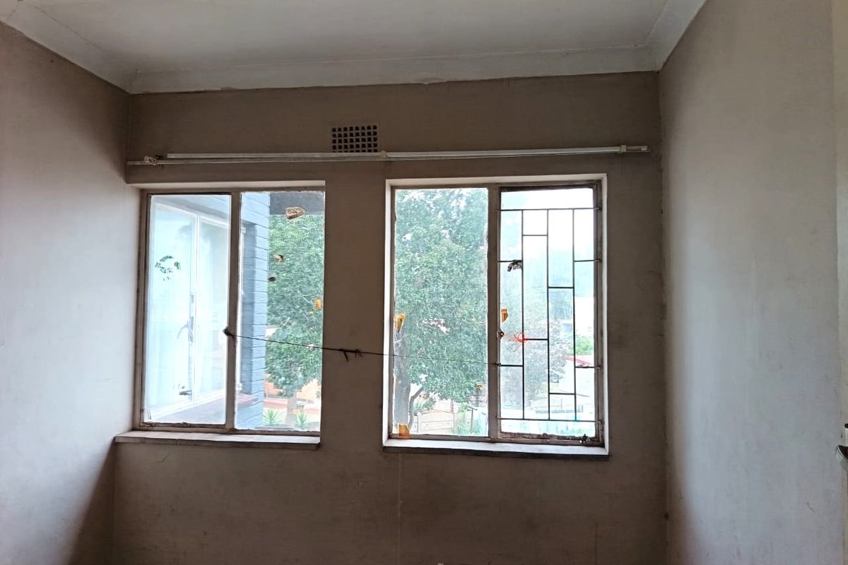 An empty bedroom featuring dual windows with security bars, ventilation grille, and curtain rail, requiring cosmetic updates to the walls and window frames.