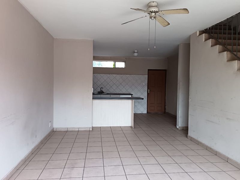 An open-plan living and kitchen area featuring tiled flooring, a ceiling fan, and a staircase, presenting a blank canvas for renovation.