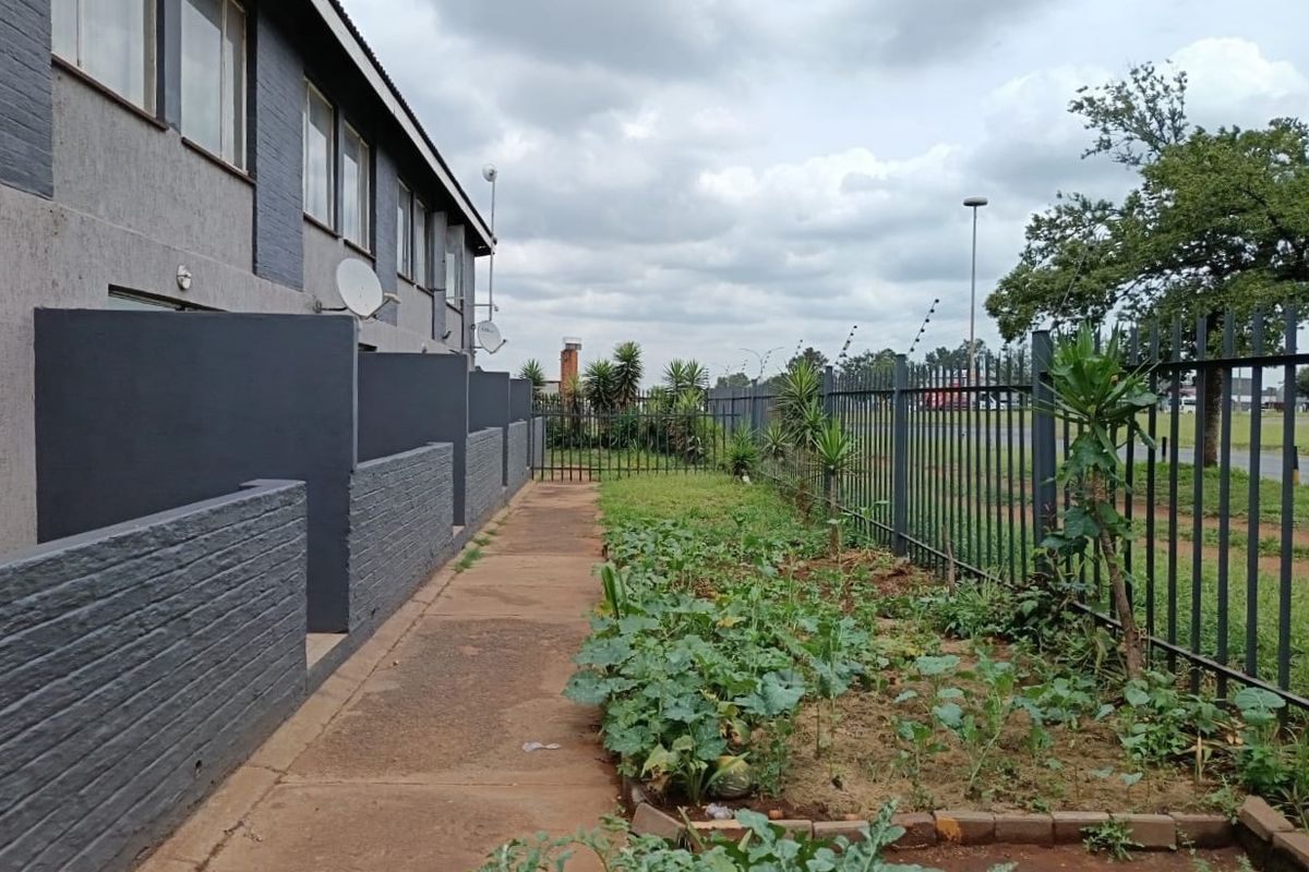 Rear exterior view of the flat featuring a paved walkway, brick privacy walls, and metal security fencing bordering the garden area.