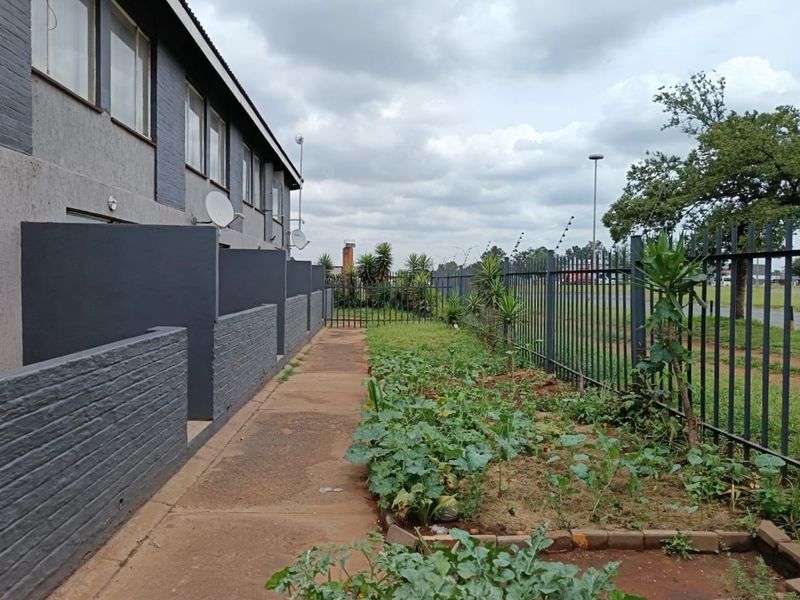 Rear exterior view of the flat featuring a paved walkway, brick privacy walls, and metal security fencing bordering the garden area.