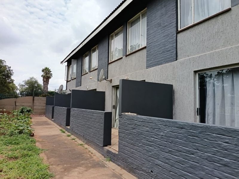 Rear view of a multi-unit flat complex featuring a paved walkway, private patio partitions, and a two-tone painted brick facade.