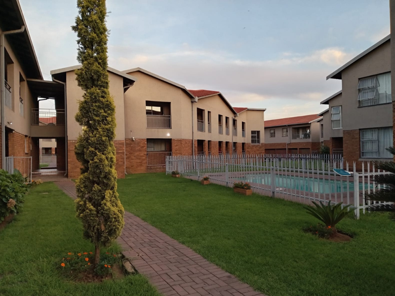 A communal pool area within a residential flat complex, featuring brick-paved pathways, landscaped lawns, and multi-story apartment buildings with balconies.