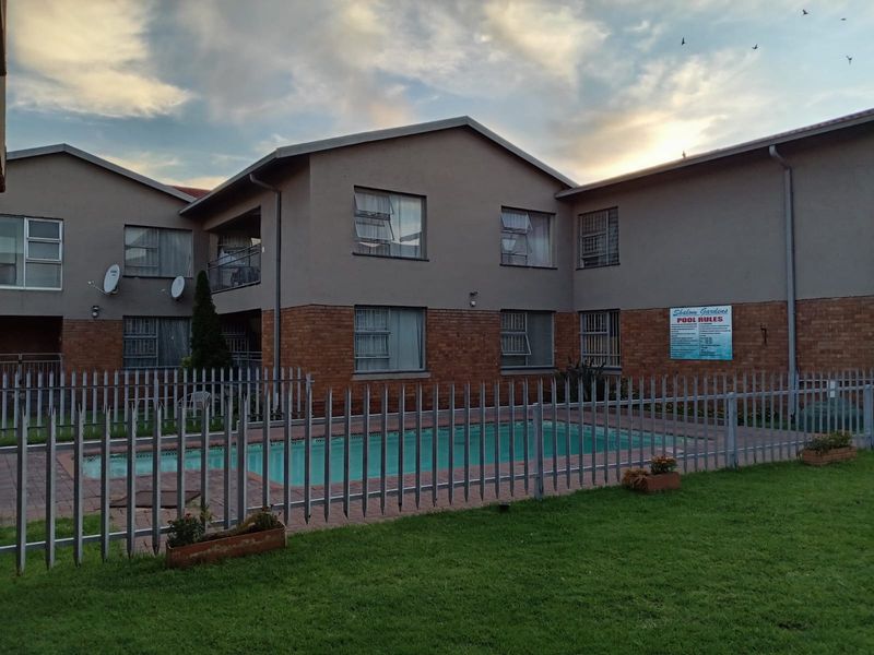 A communal swimming pool area featuring a paved surround, enclosed by metal security fencing, set against a multi-story residential building with brick and plaster finishes.