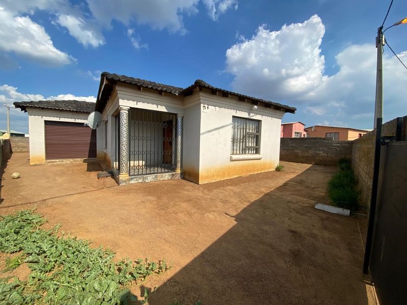Front view of a single-story house featuring a tiled roof, attached garage, and security features, set on an unpaved plot with perimeter walling.