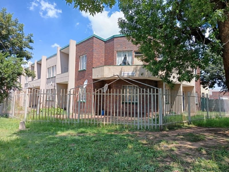 Exterior view of a face brick apartment complex featuring a secure metal perimeter fence and a paved front entrance area.