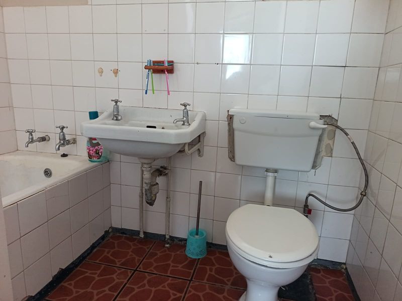 A dated bathroom featuring a wall-mounted basin, toilet, and bathtub with tiled finishes, requiring renovation to address worn fixtures and missing wall tiles.
