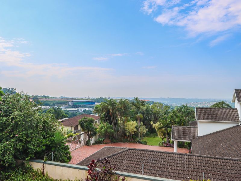 Elevated view overlooking the property's tiled roof, paved driveway, and surrounding landscaped garden with distant suburban scenery.