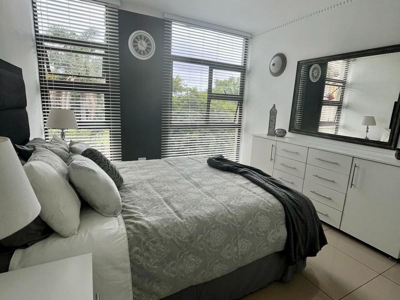 A well-maintained bedroom featuring large windows with venetian blinds, neutral tiled flooring, and a modern ceiling light fixture.