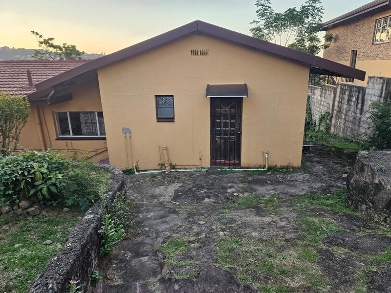 Rear exterior view of the cottage featuring a security gate, awning, and concrete paved area with surrounding retaining walls and garden space.