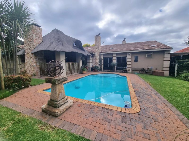 Outdoor entertainment area featuring a swimming pool, brick-paved surround, and a thatched lapa with stone-clad pillars, set within a landscaped garden.