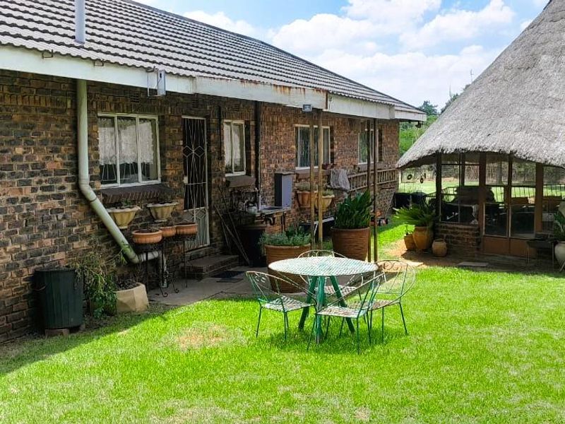 Rear view of the property featuring a face brick exterior, tiled roof, and a separate thatched lapa structure set within a garden area.
