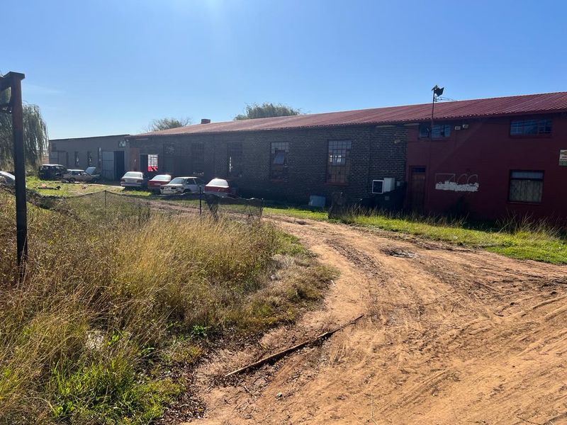 Rear view of a large industrial-style farm building featuring a corrugated metal roof and brick exterior, situated on a dirt access road.