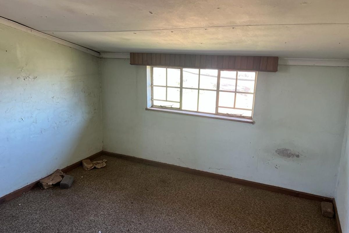 An empty bedroom featuring a sloped ceiling, carpeted flooring, and a window with security bars, requiring significant cosmetic refurbishment and wall repairs.