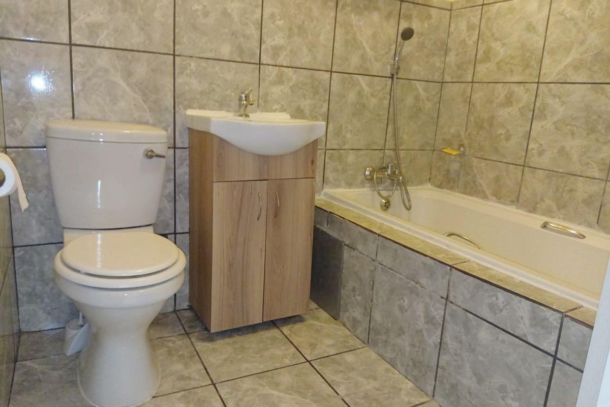 A functional bathroom featuring a bathtub with shower mixer, a vanity cabinet with washbasin, and a toilet, finished with neutral wall and floor tiles.