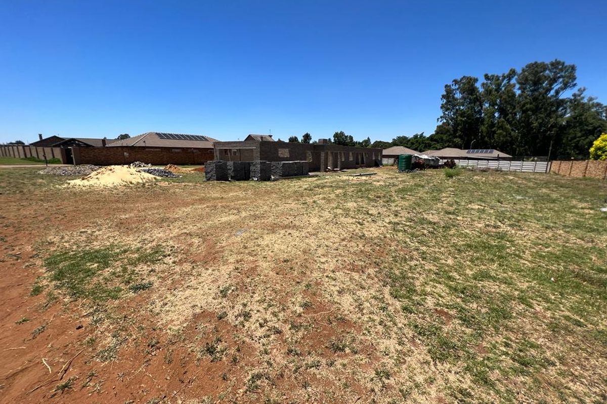A vacant residential plot featuring an ongoing brick construction project, perimeter fencing, and a water tank under a clear blue sky.