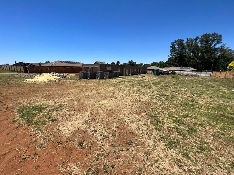 A vacant residential plot featuring an ongoing brick construction project, perimeter fencing, and a water tank under a clear blue sky.