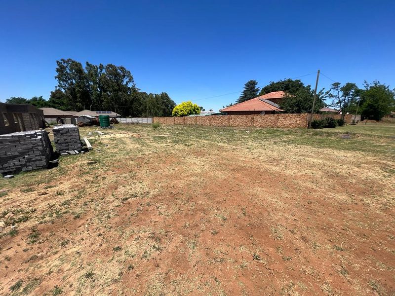 A vacant residential plot featuring a brick boundary wall, established trees, and a water tank, offering a clear space for potential development.