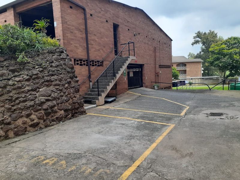 Exterior view of the apartment building featuring a brick facade, concrete staircase, and paved parking area with a security gate.