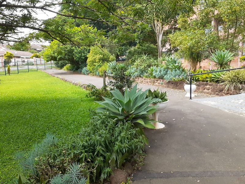 A well-maintained communal garden area featuring a paved walkway, stone retaining wall, and established landscaping within the apartment complex grounds.