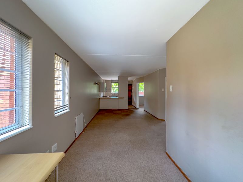 An open-plan living and kitchen area featuring neutral wall tones, carpeted flooring, and large windows with venetian blinds, offering a functional and versatile layout.