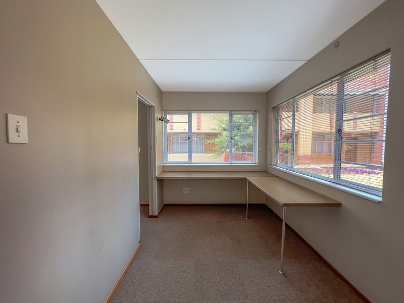 A bright, empty office space featuring built-in desk surfaces, carpet flooring, and large windows with venetian blinds.
