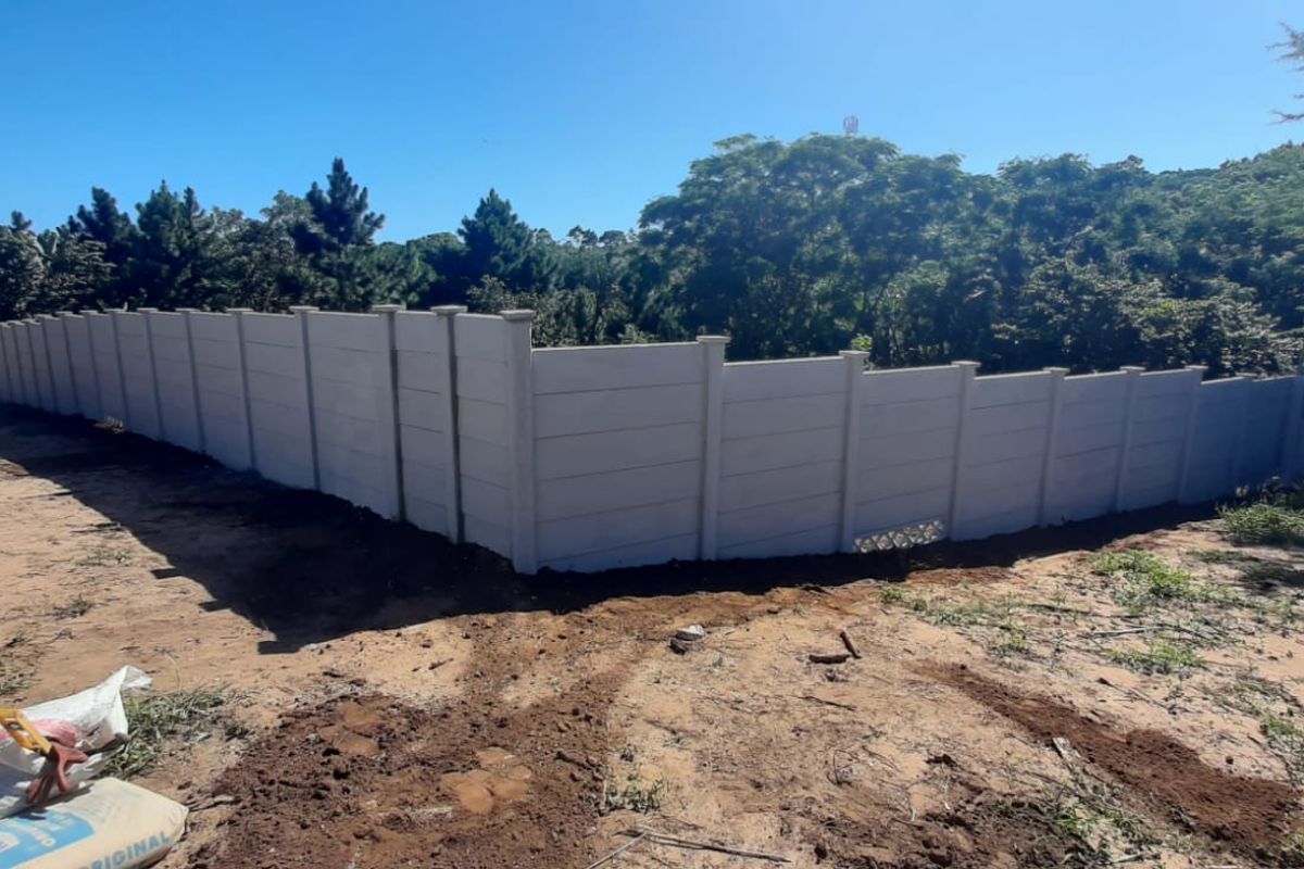 A view of a vacant residential plot featuring a newly installed precast concrete boundary wall and surrounding natural vegetation.