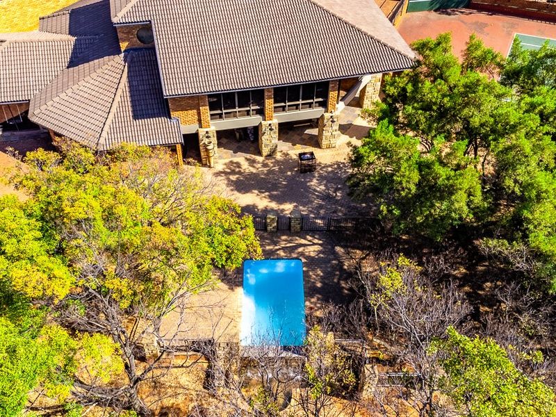 Aerial view of a residential property featuring a swimming pool, covered patio area, stone-accented architecture, and a private tennis court set within landscaped grounds.