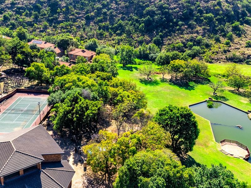 Aerial view of the property grounds featuring a private tennis court, landscaped gardens, and a scenic pond set against a hillside backdrop.