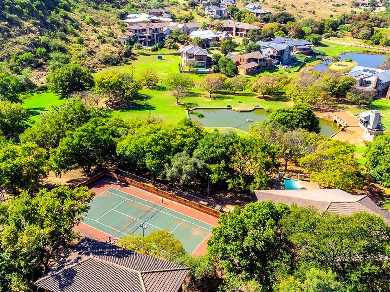 Aerial view of a residential estate featuring a private tennis court, swimming pool, landscaped gardens, and a scenic pond within a lush green environment.