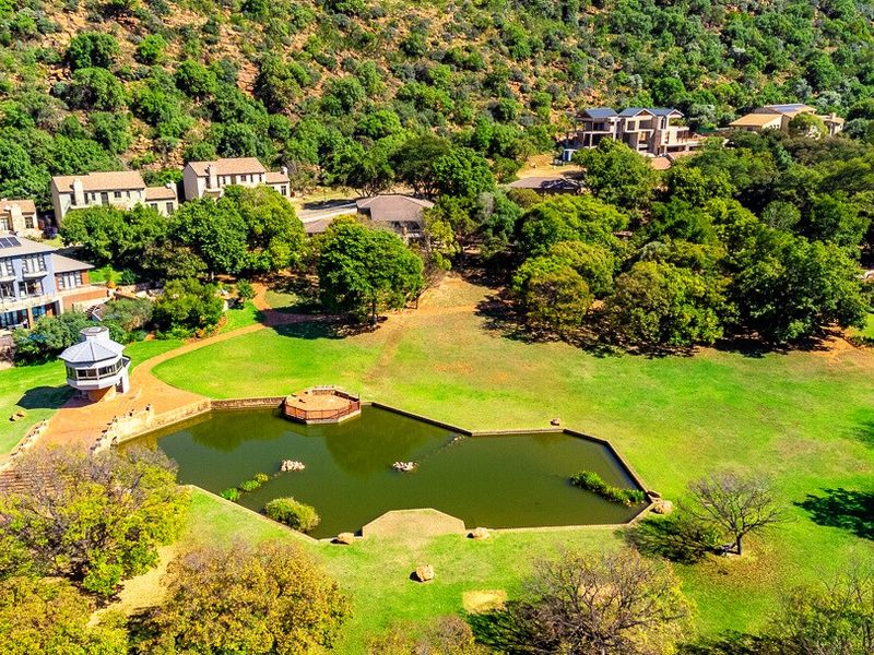 An aerial view of a residential estate featuring a large landscaped garden, a central pond with a wooden deck, and a gazebo.