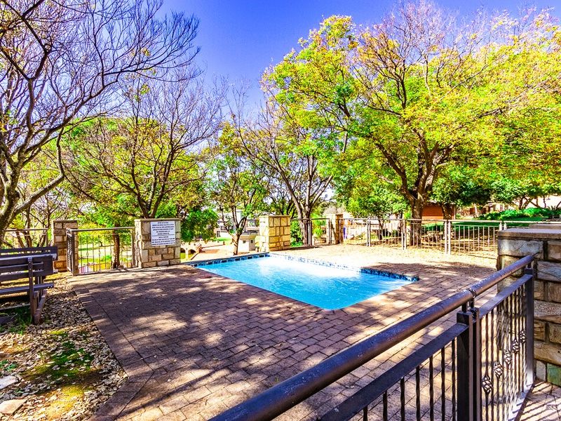 Outdoor swimming pool area featuring a paved deck, stone masonry pillars, and metal perimeter fencing surrounded by mature trees.
