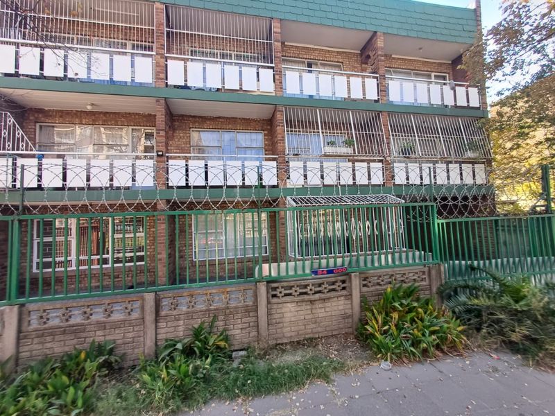 Front elevation of a multi-story brick apartment building featuring balconies with security bars and a perimeter fence with razor wire.