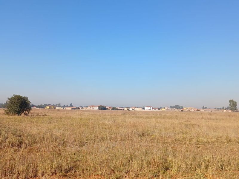 A wide-angle view of a vacant residential plot featuring dry grass and distant neighborhood housing.