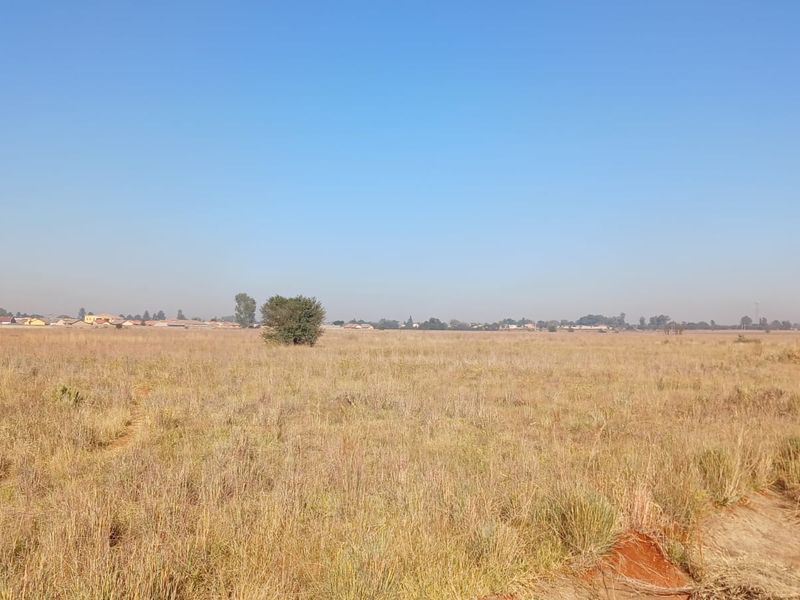 A wide-angle view of a vacant residential plot featuring dry grass and open landscape with distant neighborhood buildings visible on the horizon.