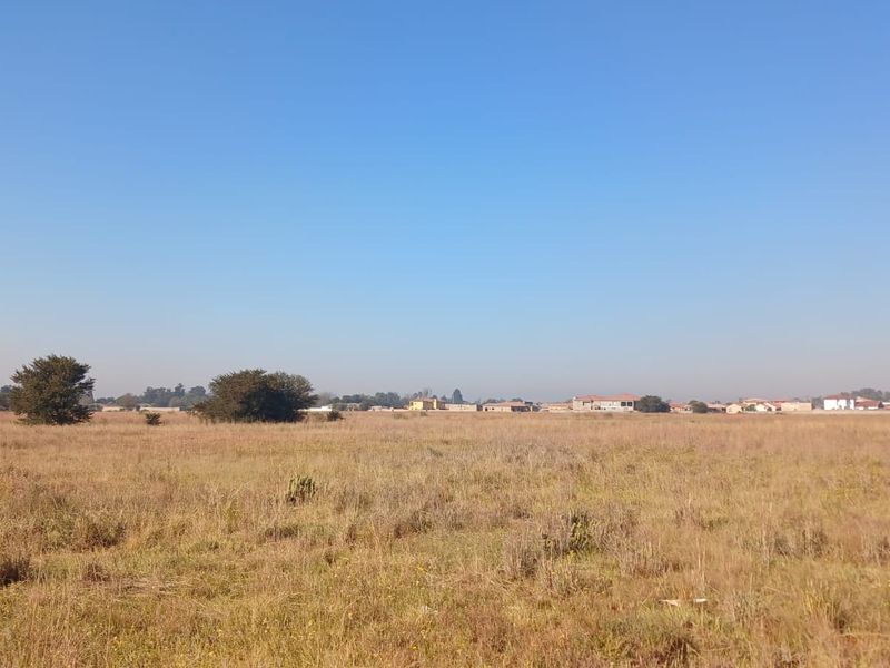 A wide-angle view of a vacant residential plot featuring dry grass and distant neighborhood buildings under a clear blue sky.