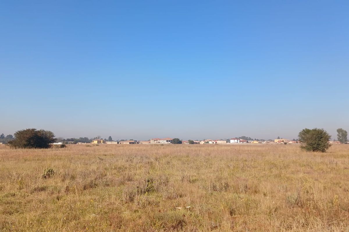 A wide-angle view of a vacant residential plot featuring dry grassland and distant neighborhood buildings under a clear blue sky.