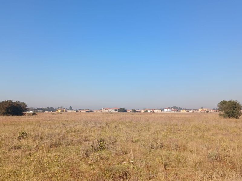A wide-angle view of a vacant residential plot featuring dry grassland and distant neighborhood buildings under a clear blue sky.