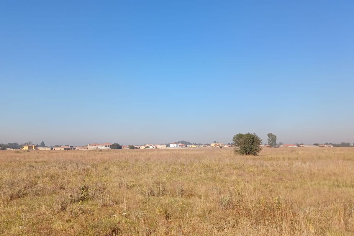 A wide-angle view of a vacant residential plot featuring natural dry grass and surrounding neighborhood landscape.
