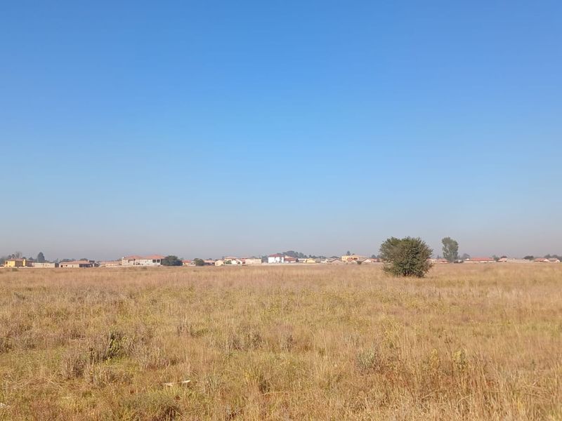 A wide-angle view of a vacant residential plot featuring natural dry grass and surrounding neighborhood landscape.