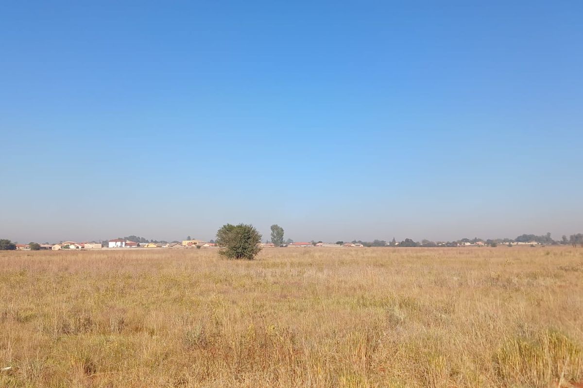 A wide-angle view of a vacant residential plot featuring dry grass and distant neighborhood housing.