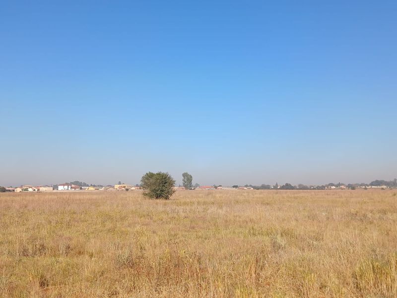 A wide-angle view of a vacant residential plot featuring dry grass and distant neighborhood housing.